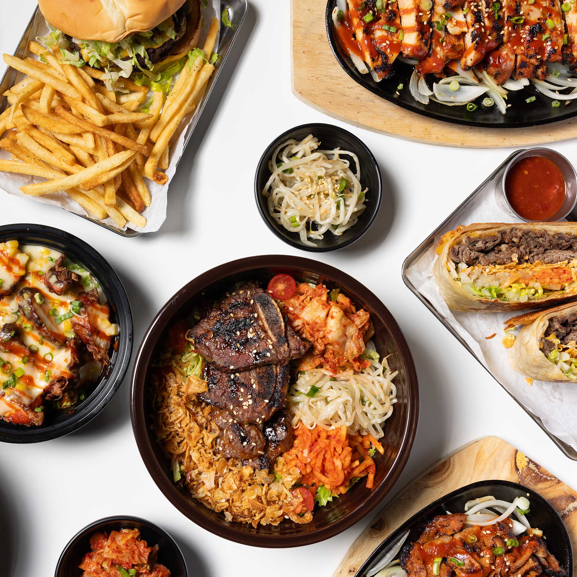 a variety of the menu plated together and shot from above, with burgers, crunchwraps, banchan, kimchi fried rice, and spicy chicken plate all in frame