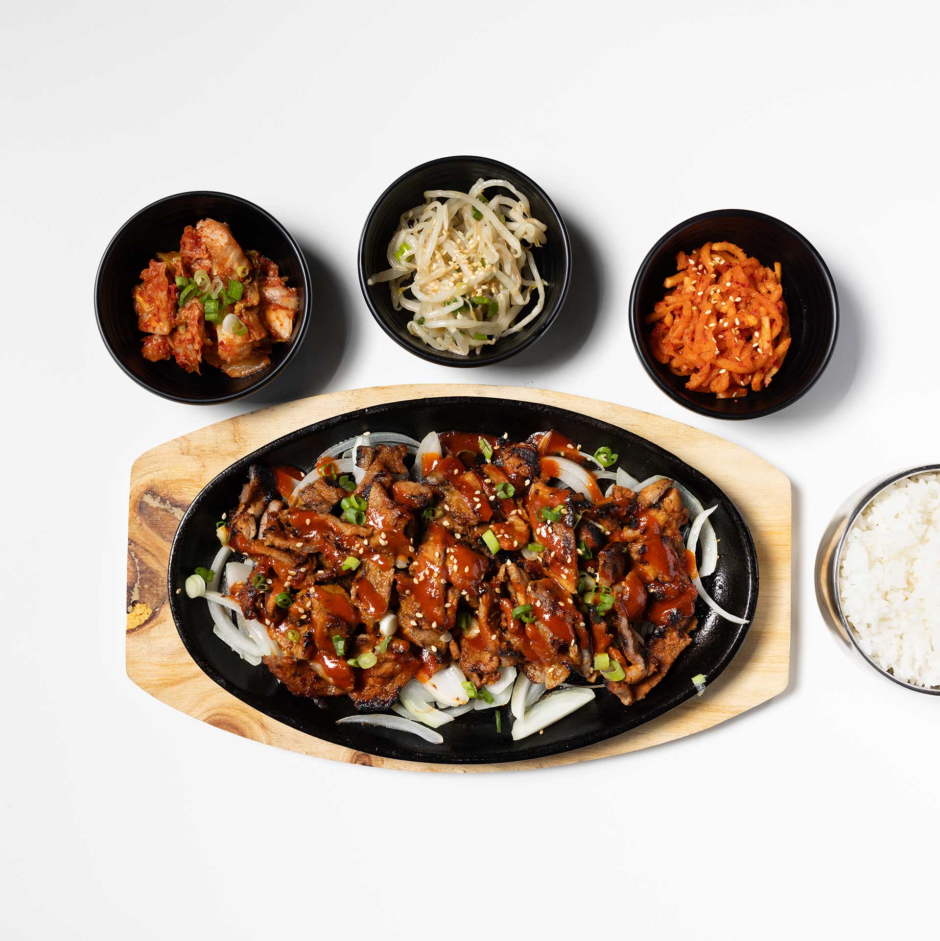 skillet of galbi short ribs on a wood plate with sides of kimchi, sprouts, and rice in small bowls surrounding it